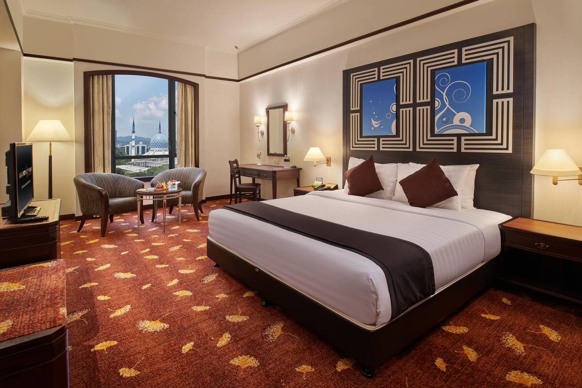 Ein Hotelzimmer mit einem großen Bett, einem Schreibtisch, einem Sessel, einem Fernseher und Blick auf eine Stadtlandschaft.