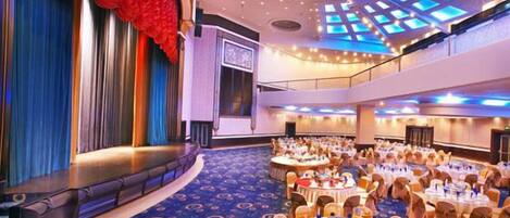 A large hall with a stage, multiple round tables, and a patterned carpet.