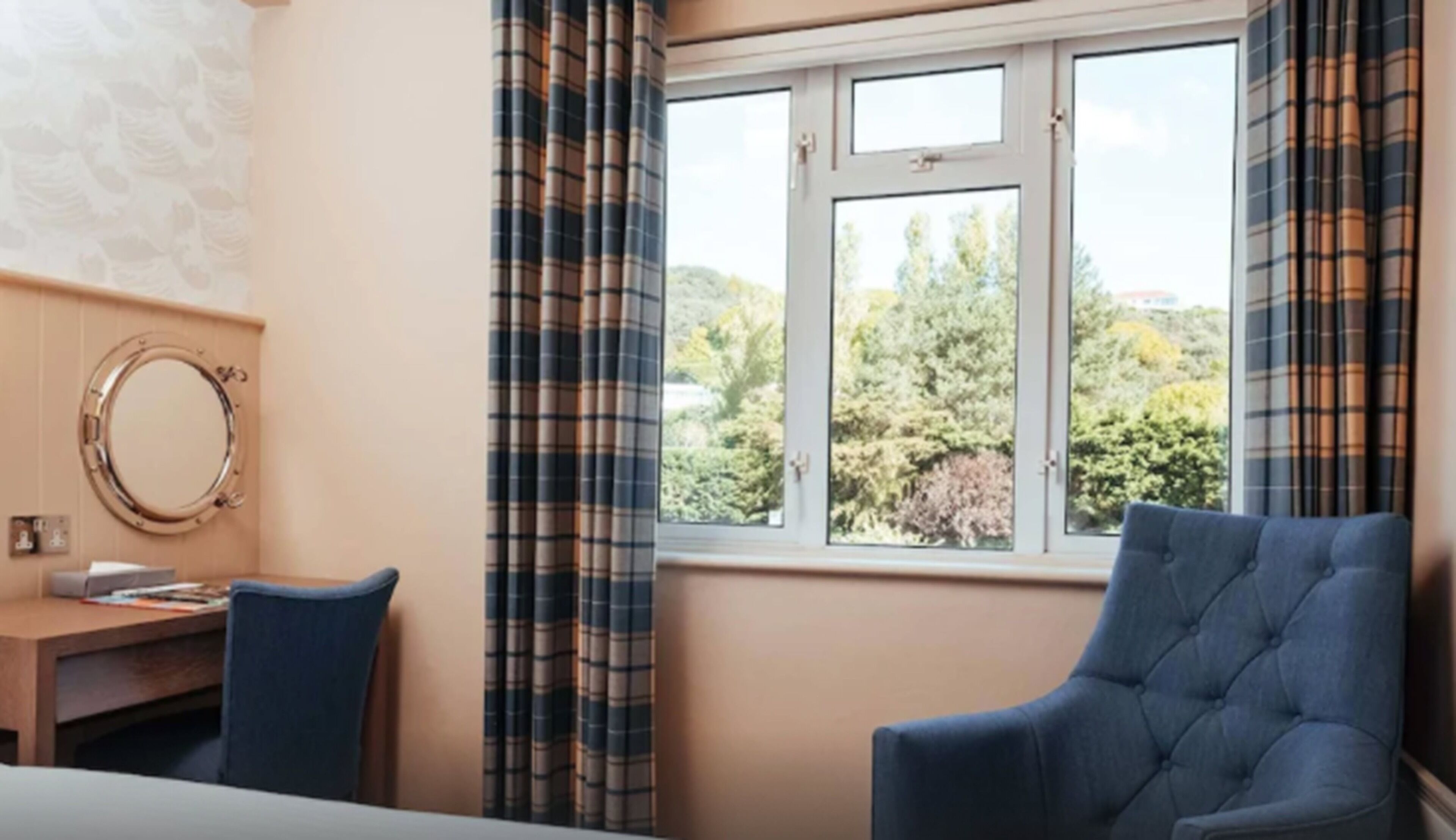 A room with a large window, a round mirror, a blue chair, and a wooden desk.