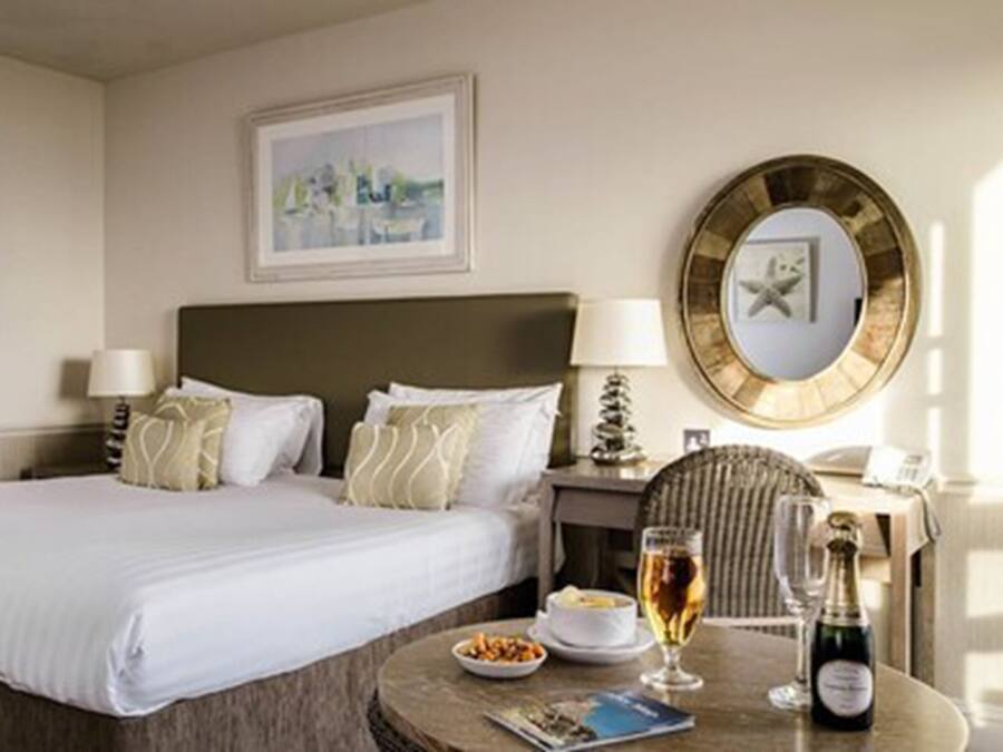 Una habitación de hotel con una cama grande, dos mesitas de noche con lámparas, un espejo redondo, una pequeña mesa redonda con una copa de vino y una botella, y un cuadro en la pared.