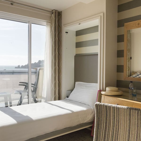 A hotel room with a bed, a chair, a small table, a mirror, and a view of the sea. - The Golden Sands (St. Brelade)