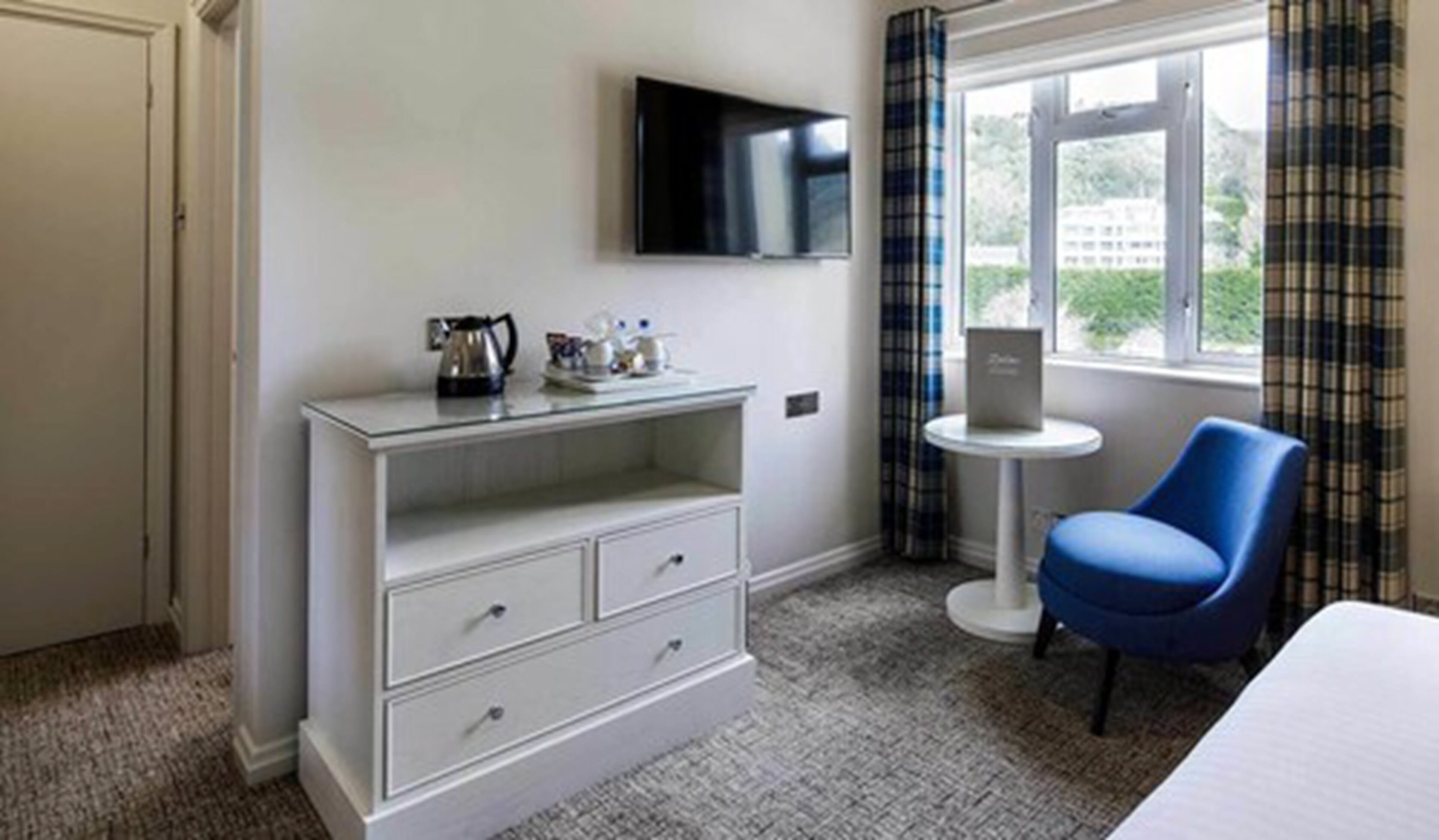 A hotel room with a white dresser, a blue chair, a small table with a laptop, and a TV mounted on the wall.