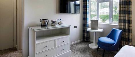 A hotel room with a white dresser, a blue chair, a small table with a laptop, and a TV mounted on the wall.