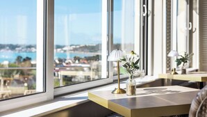 Et værelse med stort vindue der giver udsigt over byen, et lille bord med lampe og en vase med blomster, samt en stol.