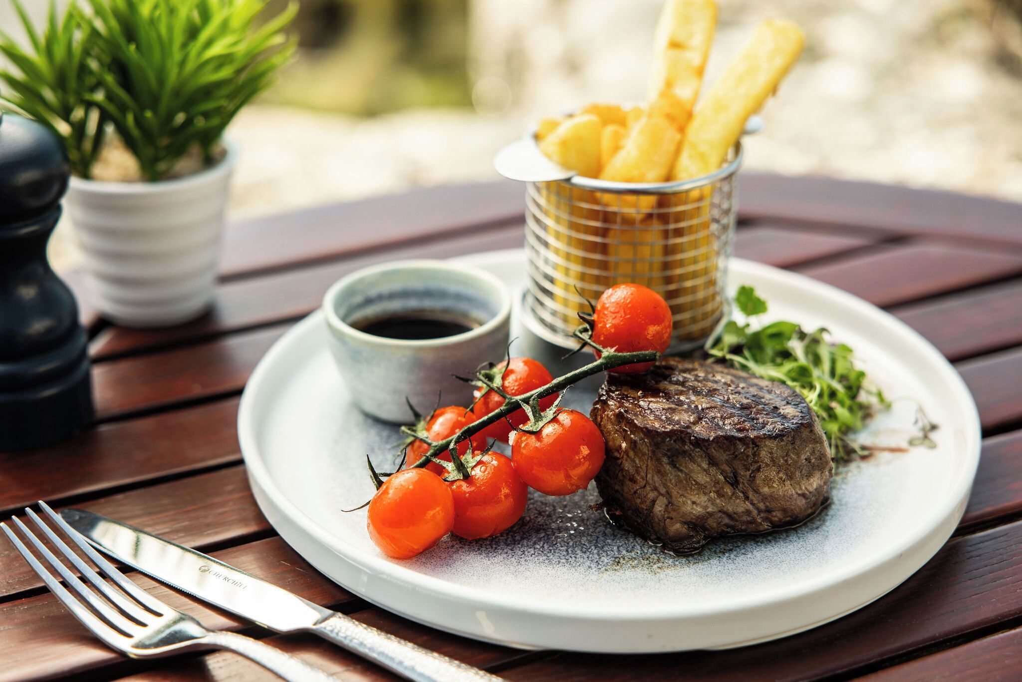 A plate with steak, cherry tomatoes, fries, and a small cup of sauce on a wooden table.