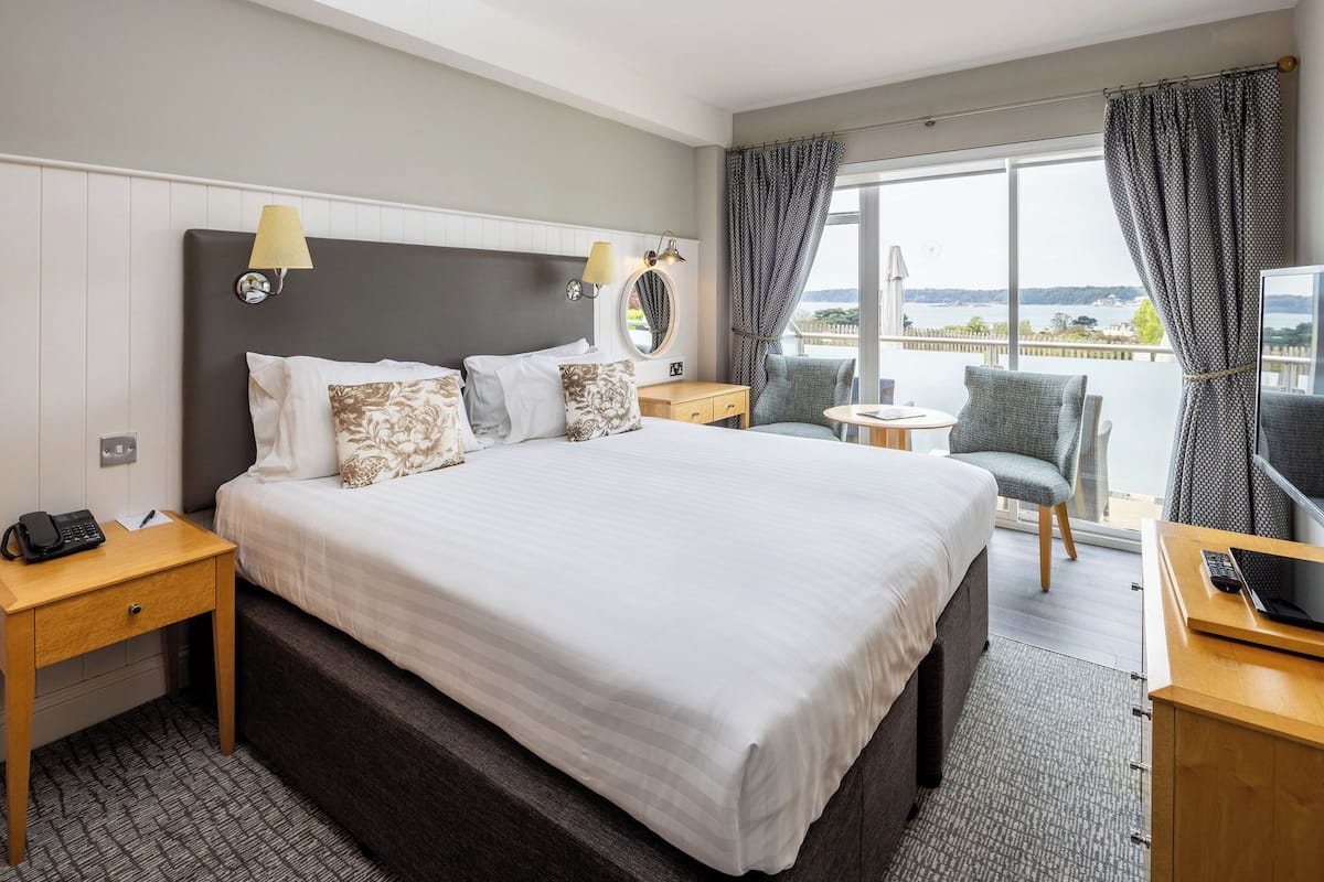 A hotel room with a large bed, bedside lamps, a small desk with a phone, a chair, and a view of the sea.