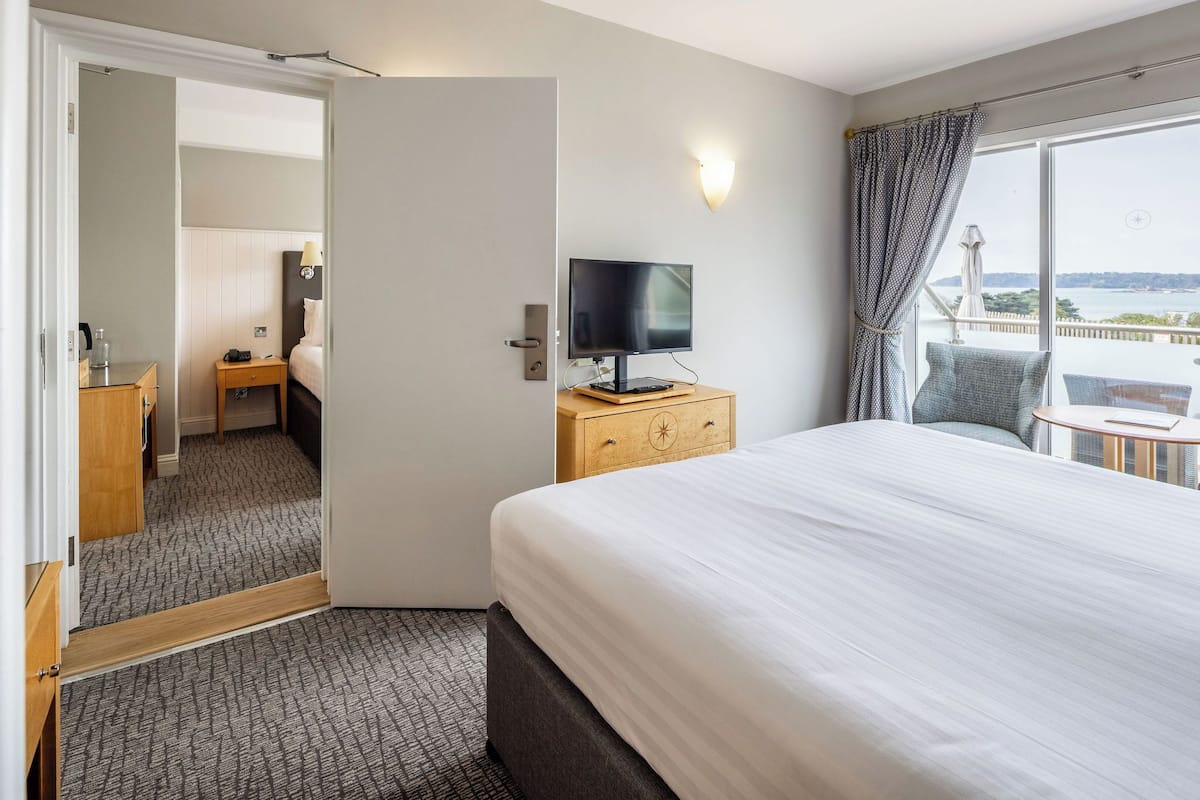 A hotel room with a bed, a TV on a wooden nightstand, a desk with a lamp, a chair, and a balcony with a view.
