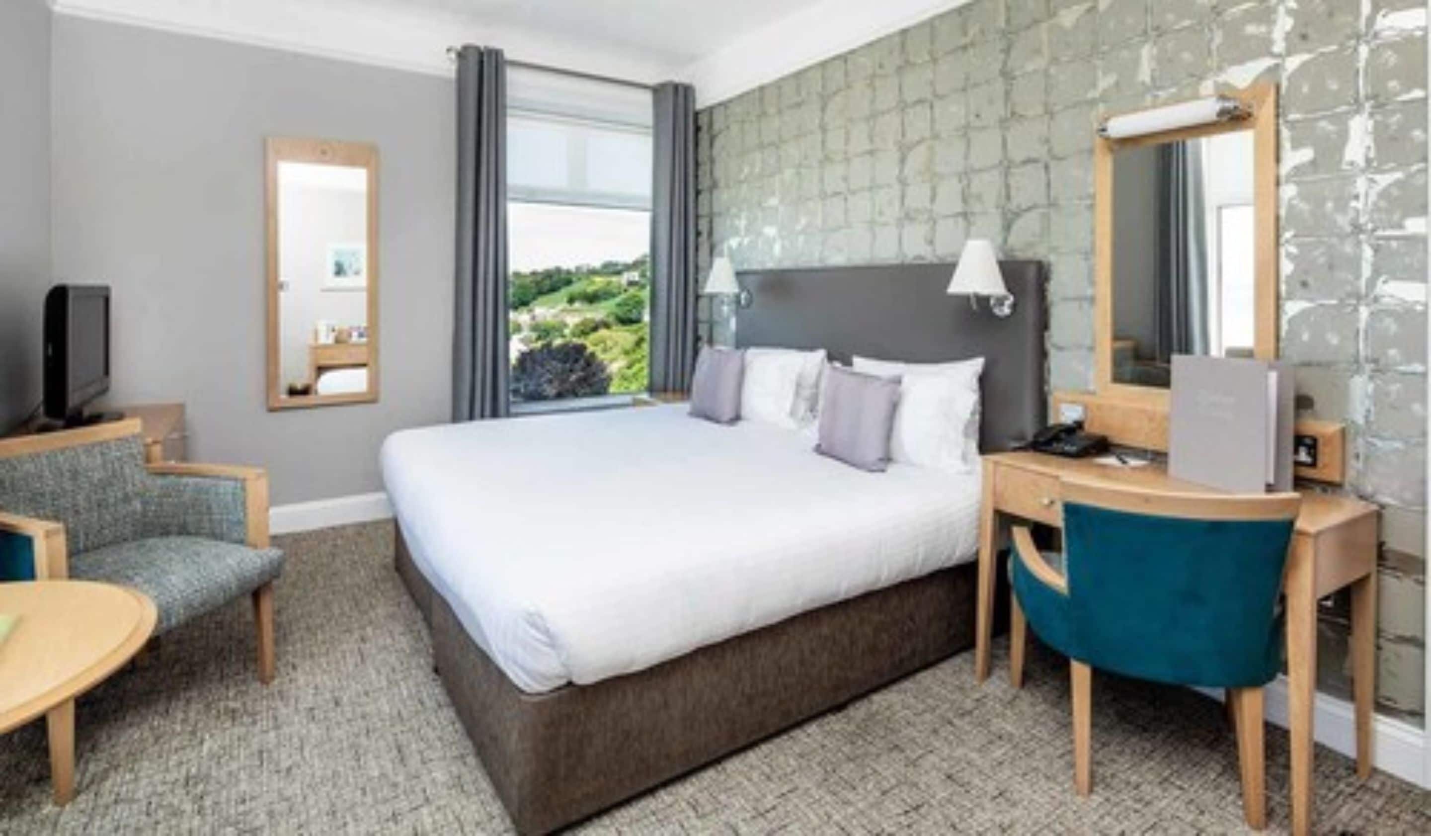 A hotel room with a large bed, a desk, a chair, a TV, and a window with a view of greenery.