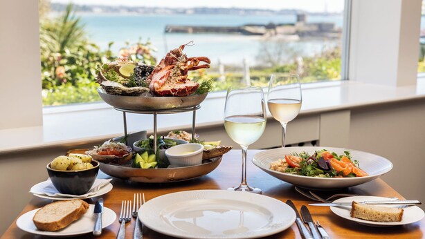 A table set with seafood, bread, and wine glasses, overlooking a scenic view of the sea and a bridge. - Somerville Hotel (St. Brelade)