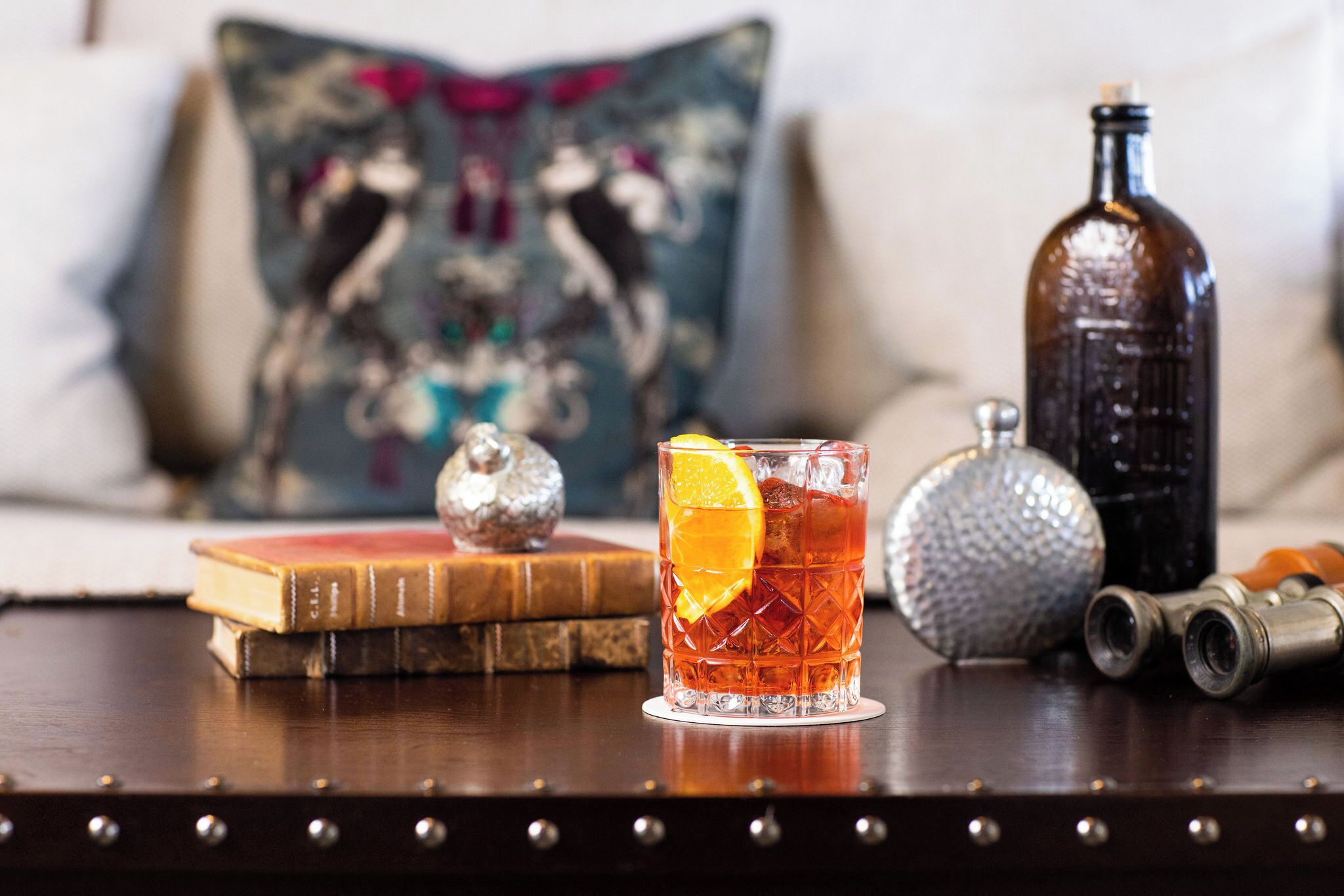 Un vaso de bebida color ámbar con una rodaja de naranja, una botella antigua, un objeto metálico decorativo y un libro sobre una superficie de madera.