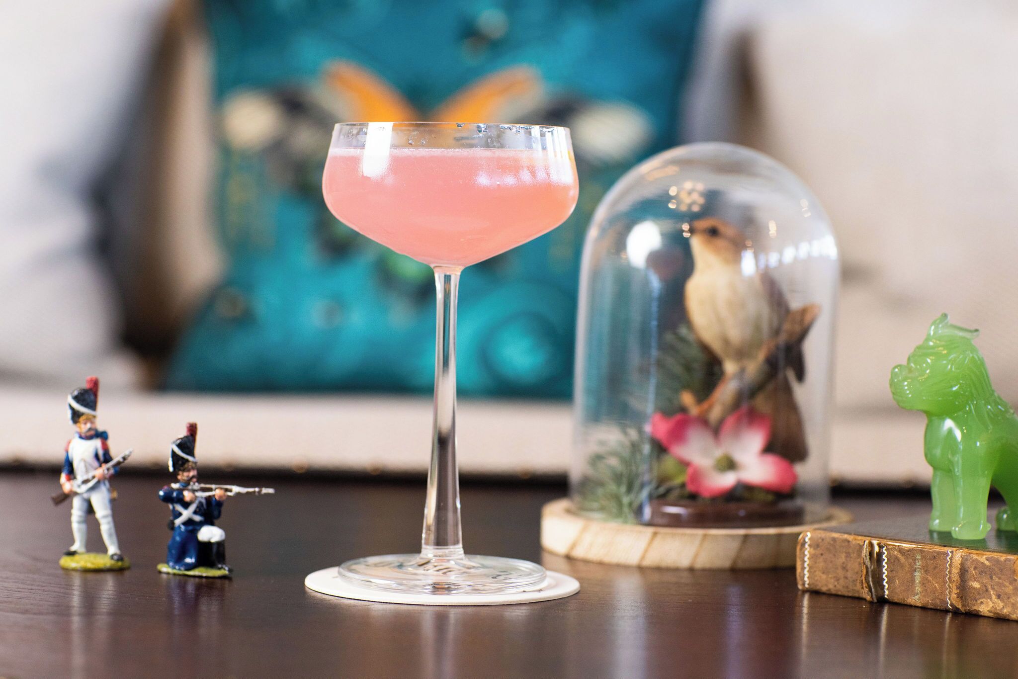 Un vaso de cóctel con una bebida rosada, miniaturas, una cúpula de cristal con un pájaro y una figurita verde sobre una superficie de madera.