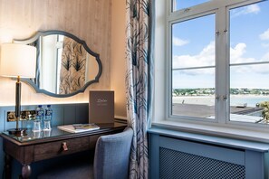 A hotel room with a desk, a mirror, a window with a view of the beach, and a thermostat.
