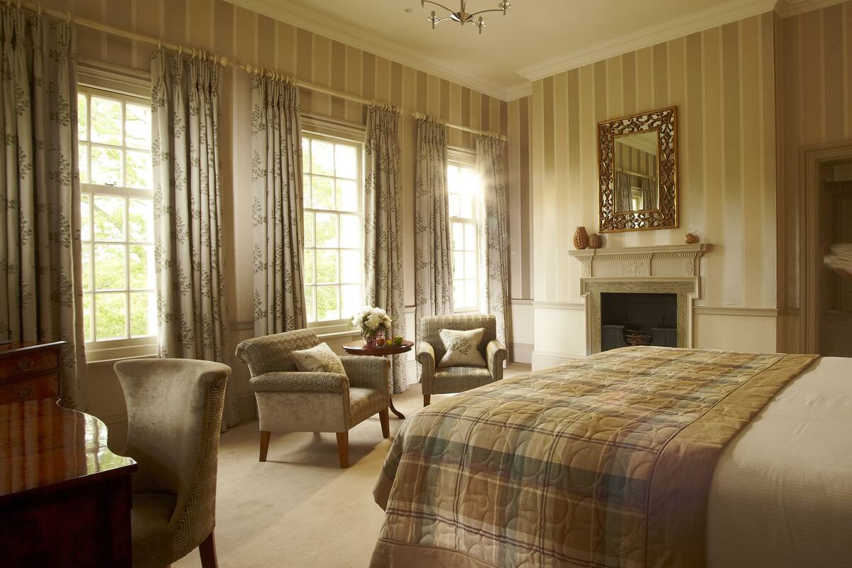 A room with a large bed, two armchairs, a fireplace, and a mirror on the wall.