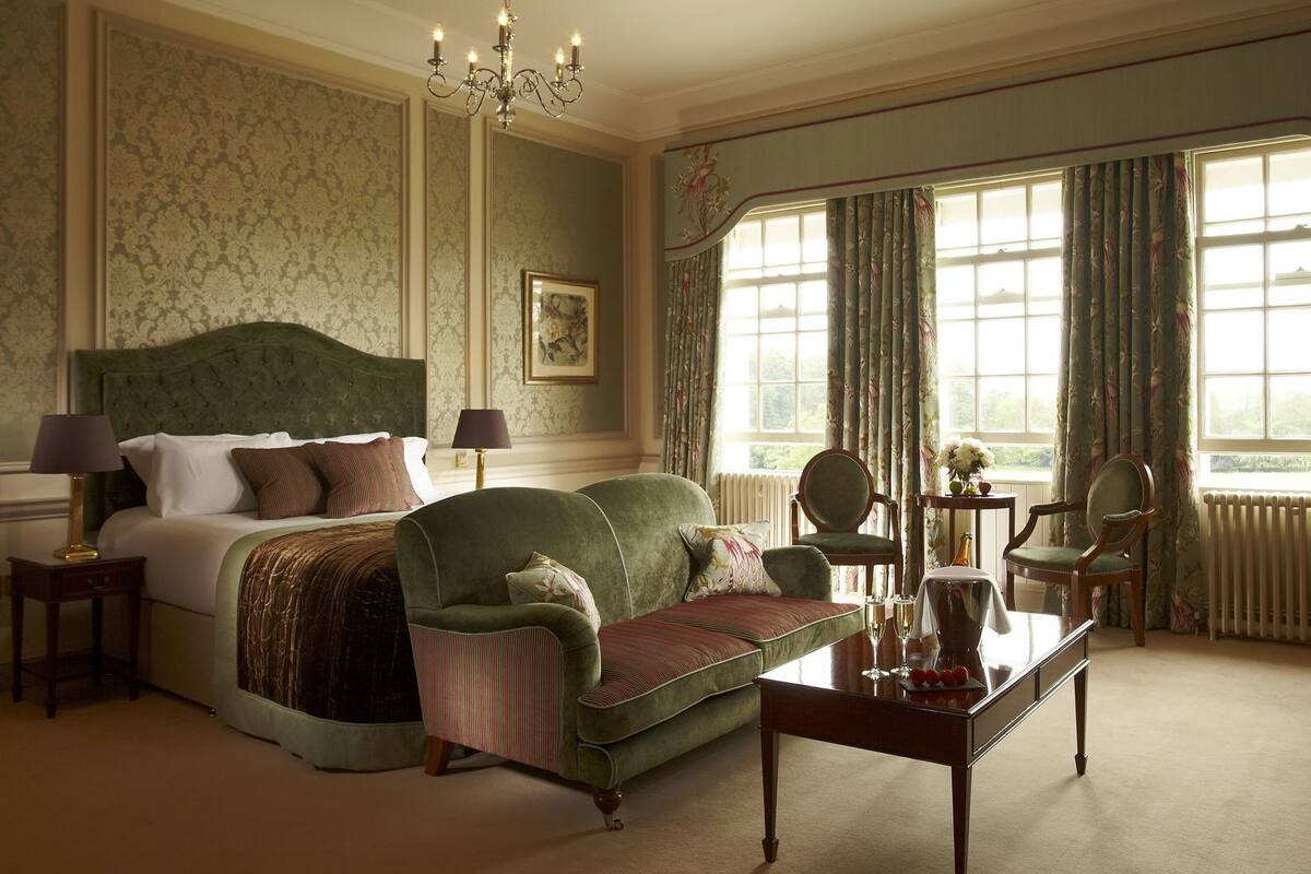 A hotel room with a bed, a green sofa, a wooden coffee table, a chandelier, and large windows with curtains.