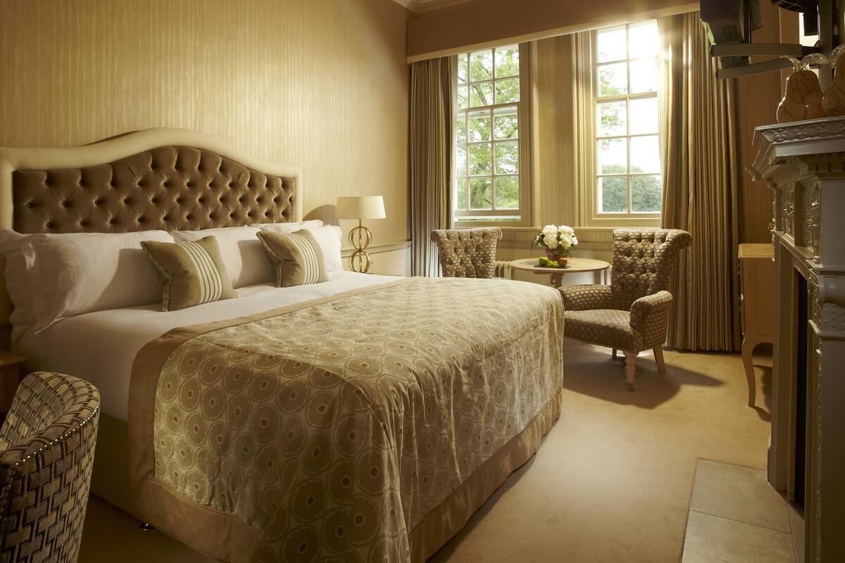 A spacious bedroom with a large bed, two armchairs, a small round table, and a fireplace.