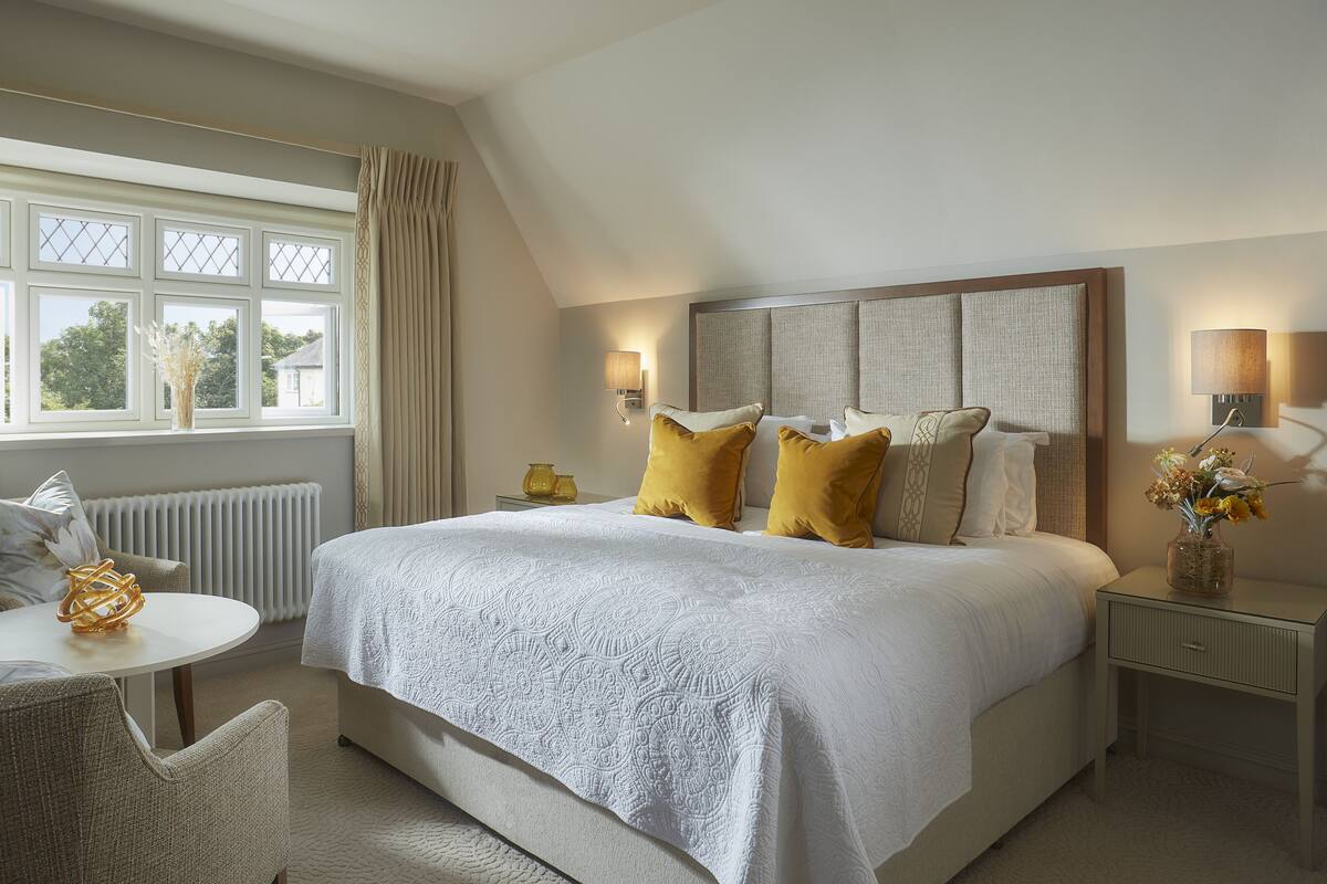 A bedroom with a large bed, a small table, a chair, a nightstand, a lamp, and a window with curtains.