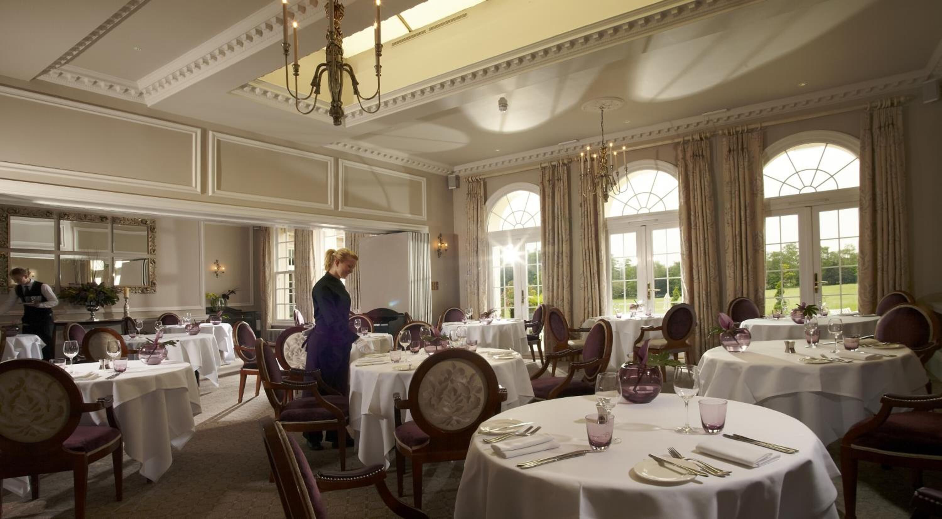 Een formele eetzaal met meerdere ronde tafels, elegante kroonluchters en grote ramen met uitzicht op het buiten.