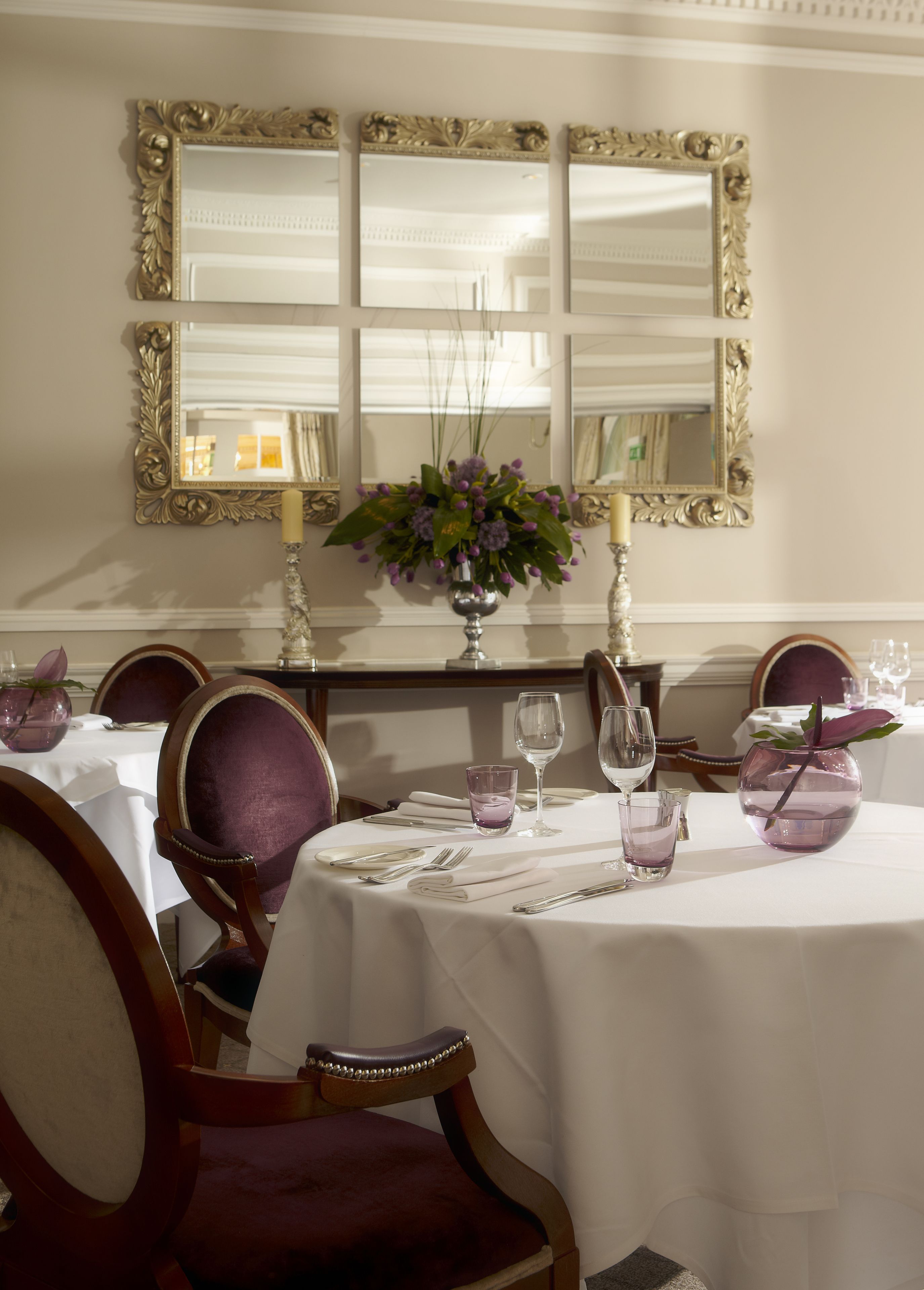 Un comedor con una mesa dispuesta para cuatro personas, rodeada de sillas, un florero con flores y velas.