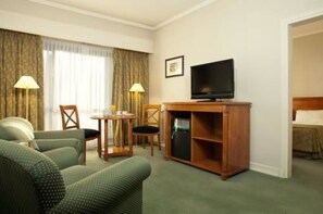 A hotel room with a green sofa, a wooden TV stand, a small table with two chairs, and a bed with a headboard.