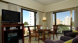 A hotel room with a television, a dining table with chairs, a sofa, and large windows offering a city view. - Austral Hotel (Comodoro Rivadavia)