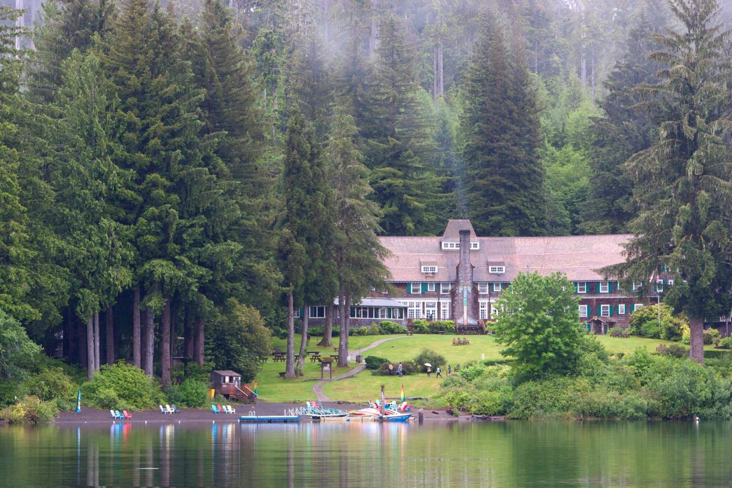 A lakeside lodge with a large building surrounded by dense forest, a dock with boats, and a clear view of the water.