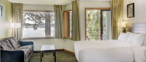 A hotel room with a bed, a sofa, a small table, and two windows offering a view of a lake and trees.