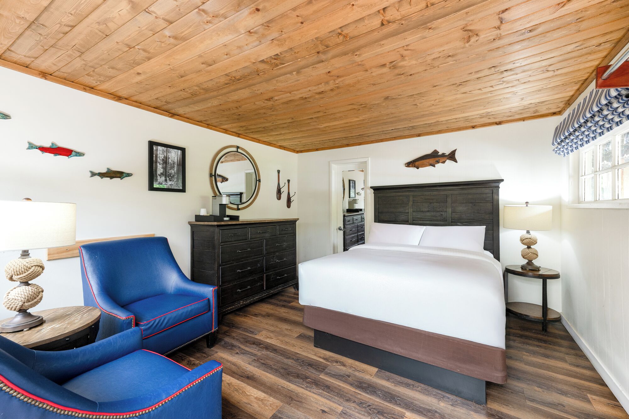 Un dormitorio con techo de madera, una cama con cabecera oscura, un sillón azul, un aparador y un espejo.
