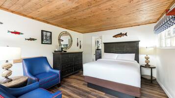 A bedroom with a wooden ceiling, a bed with a dark headboard, a blue armchair, a dresser, and a mirror.