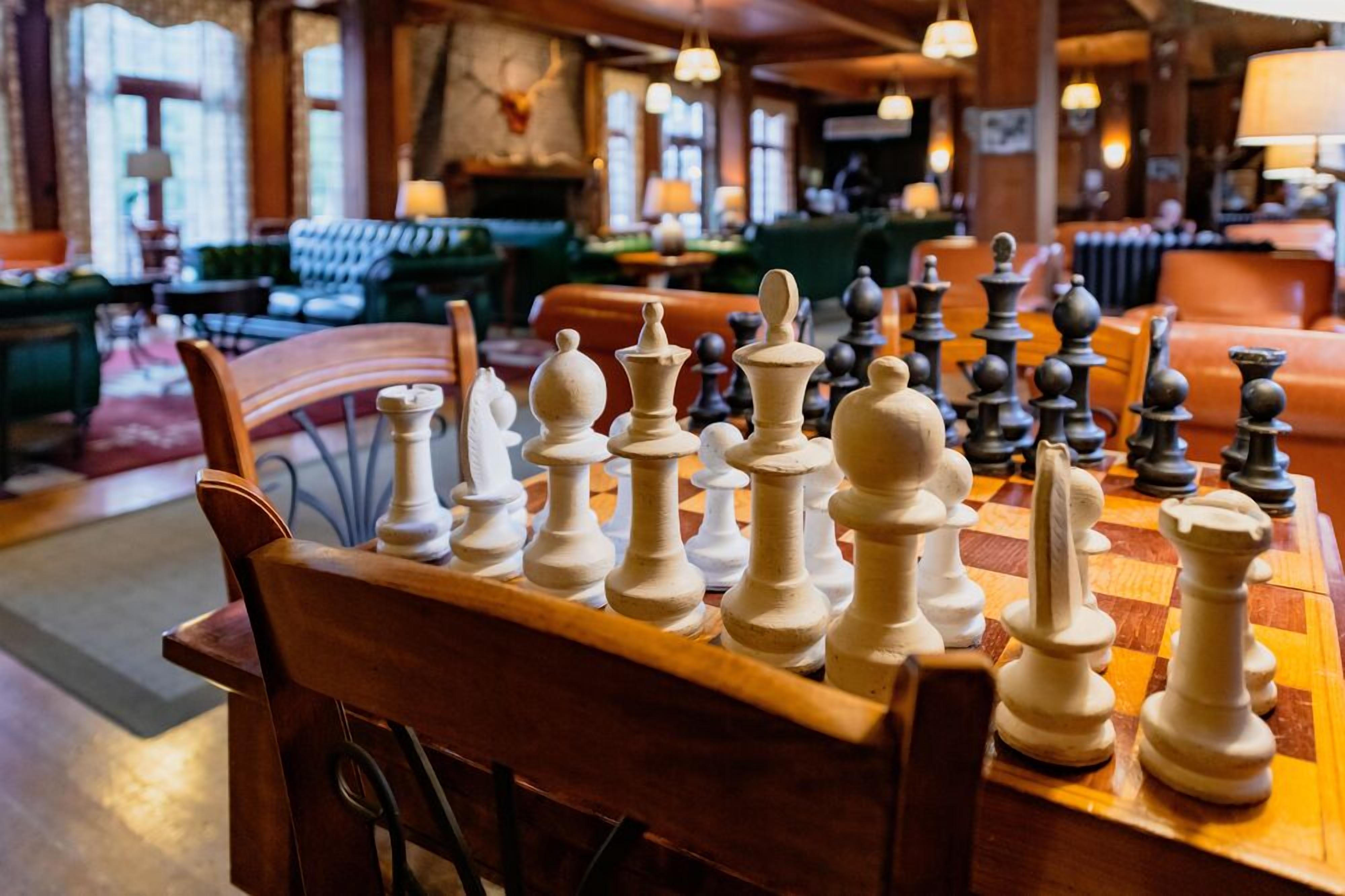 A chessboard set up on a wooden table in a cozy, well-lit room with comfortable seating and large windows.