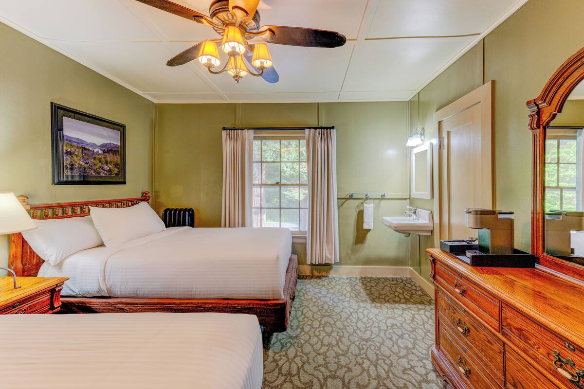 Une chambre d’hôtel avec deux lits, un ventilateur de plafond, une fenêtre avec des rideaux, une commode et un tableau encadré au mur.