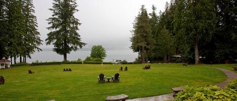 A grassy outdoor area with several wooden benches and picnic tables, surrounded by tall trees and a view of a body of water.