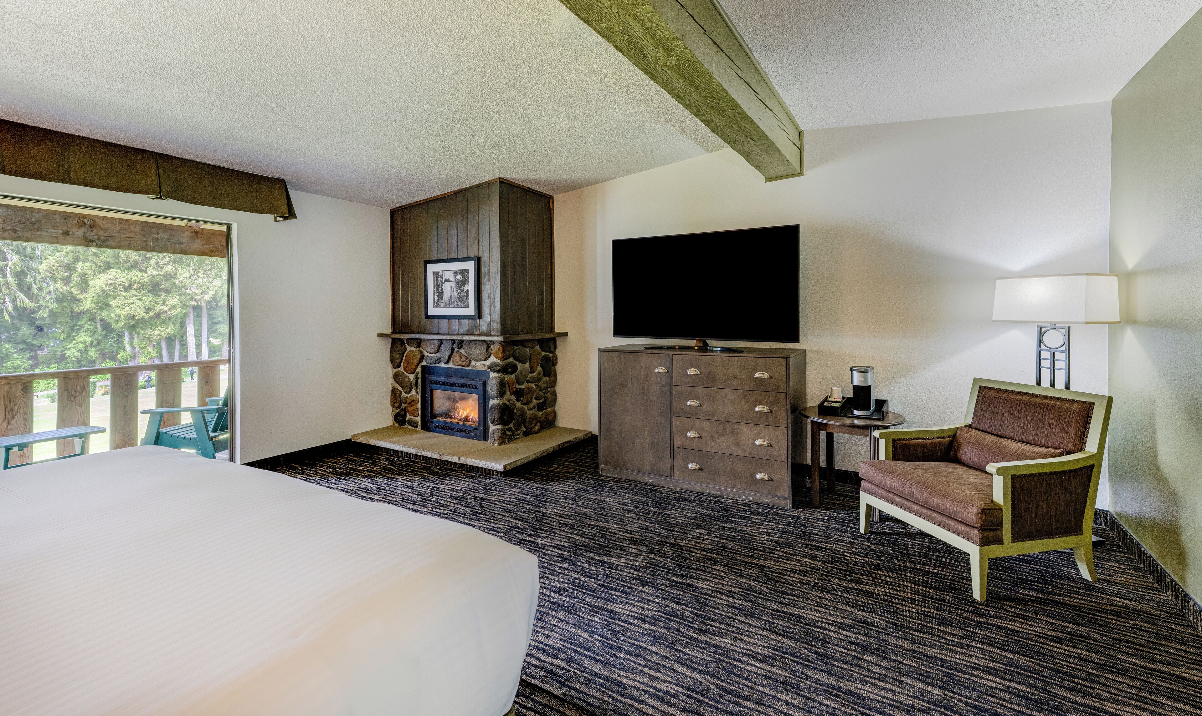 Hotelkamer met een groot bed, een stenen openhaard, een flatscreen televisie, een bureau met een lamp en uitzicht op bomen vanaf een balkon.