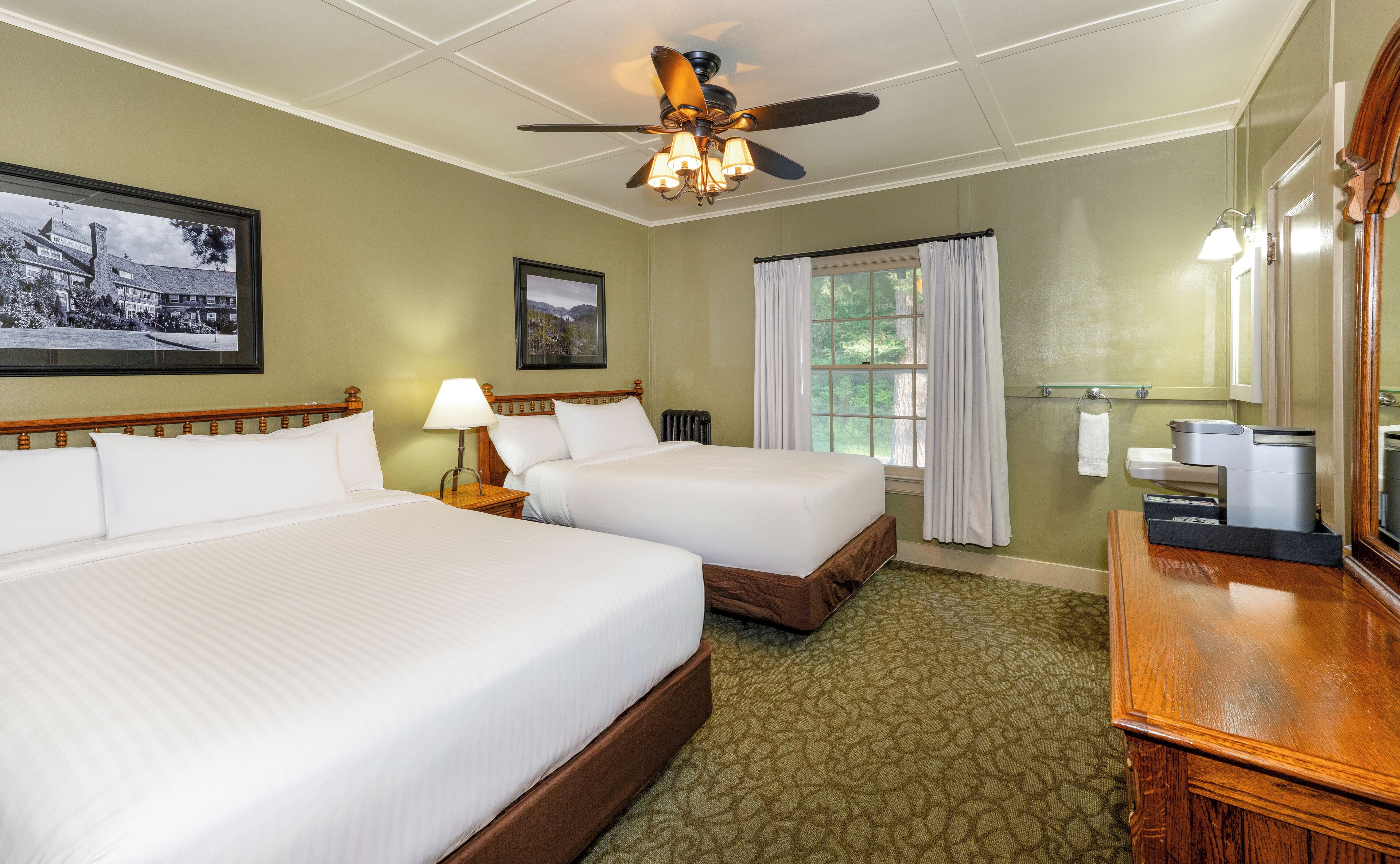 A hotel room with two beds, a ceiling fan, a framed picture, a window with curtains, a bathroom with a mirror and towel rack, and a wooden dresser.