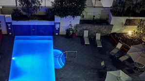 An outdoor swimming pool with a blue-lit edge, surrounded by lounge chairs and umbrellas, and a stone wall with plants. - West End Central Apartments (South Brisbane)