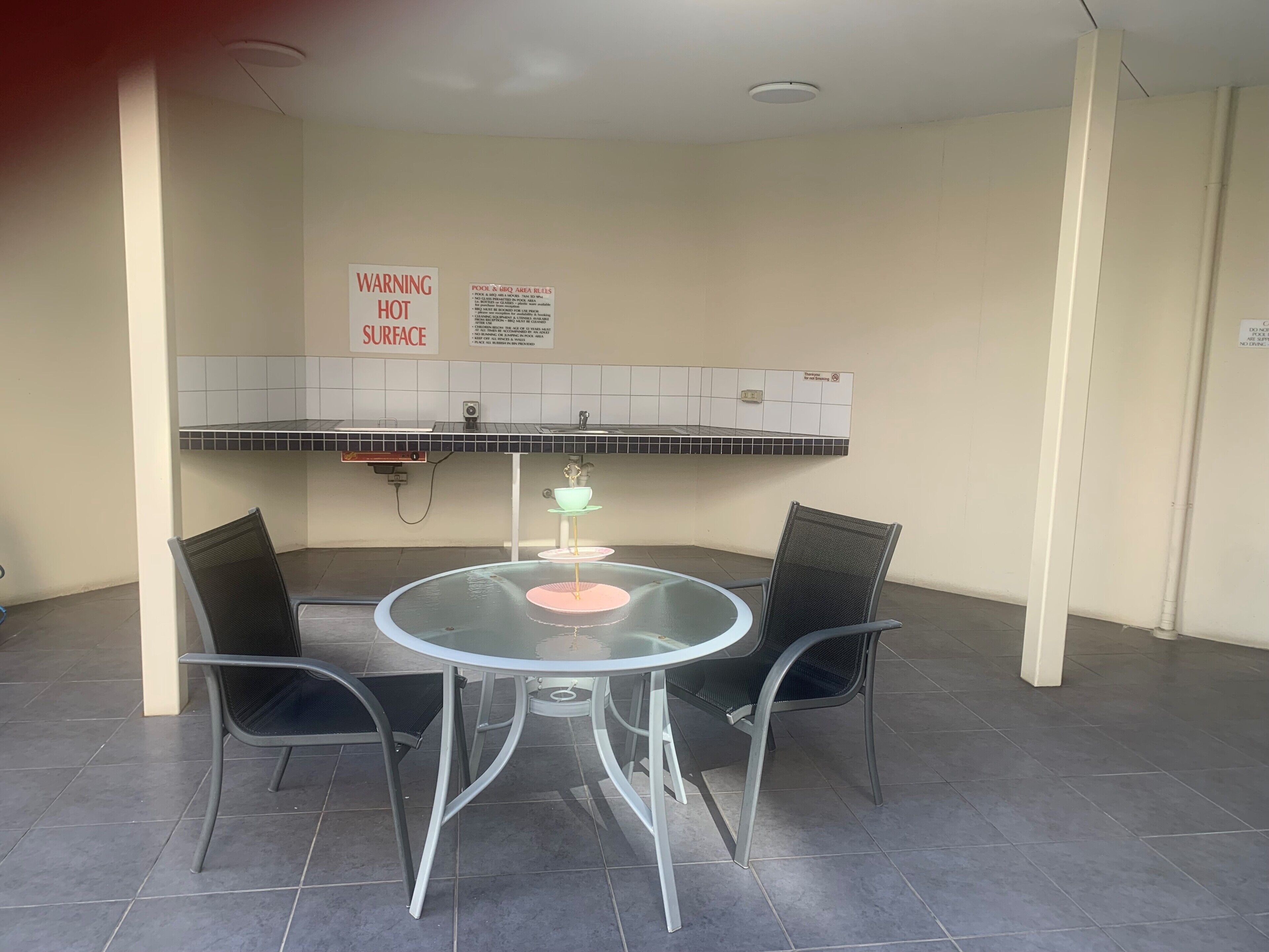 Un petit espace clos comprenant une table ronde et deux chaises, un panneau d’avertissement indiquant une surface chaude, et un pare-éclaboussures carrelé au-dessus de l’évier.