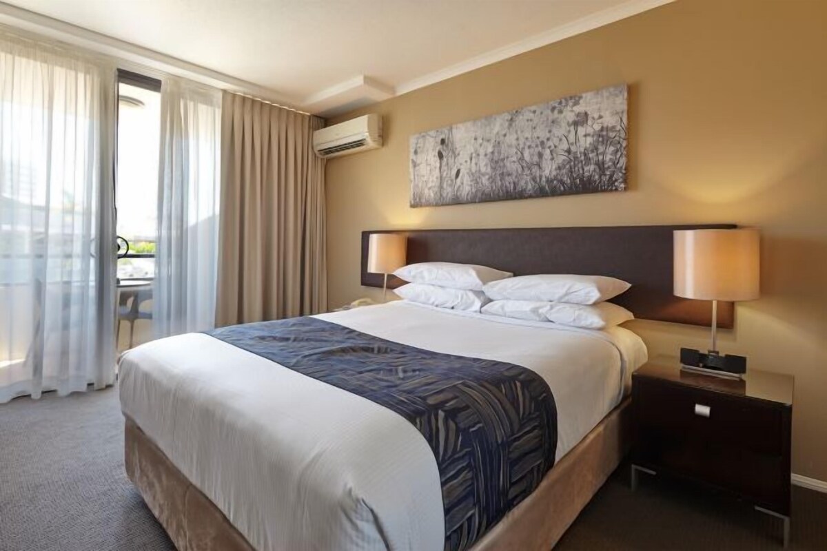 Habitación de hotel con una cama grande, dos lámparas de noche, un cuadro en la pared y vistas al exterior a través de una ventana con cortinas.