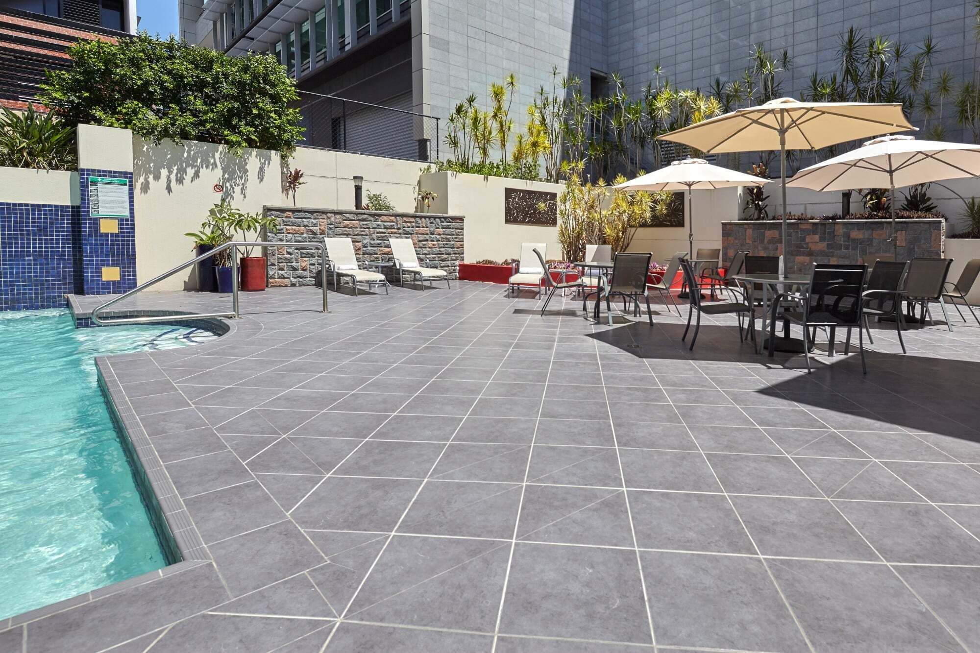 A poolside area with a tiled floor, lounge chairs, umbrellas, and a stone wall.