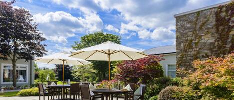 An outdoor patio with a table and chairs, surrounded by lush greenery and a stone building.