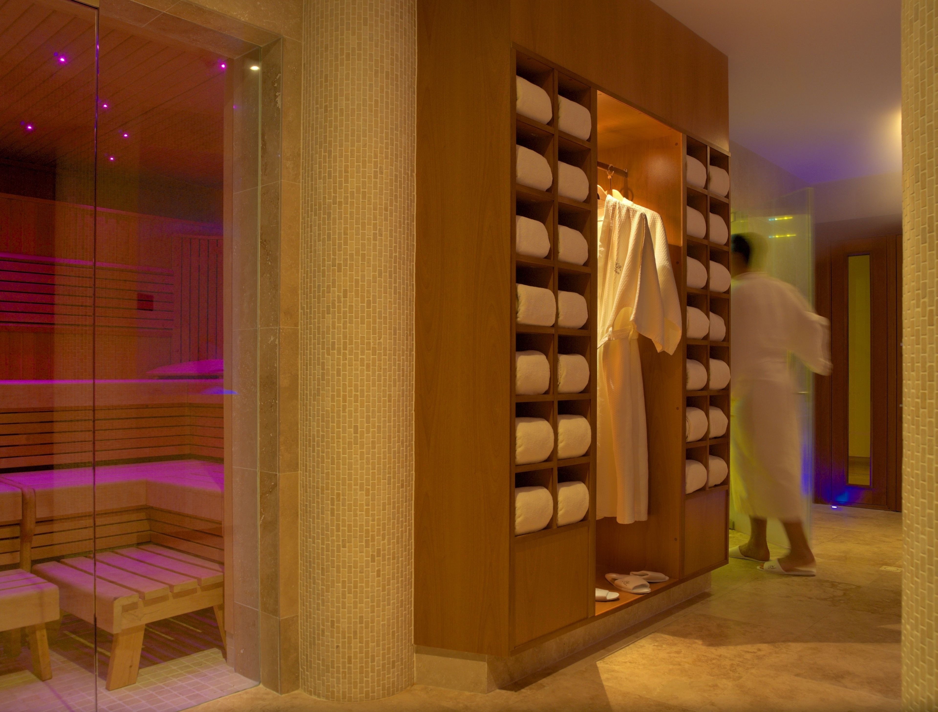 A spa room with a wooden bench, a massage table, and a wall with shelves holding towels and slippers.