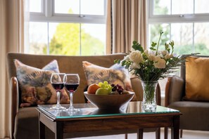 Una mesa de comedor con un frutero y dos copas de vino, un jarrón con flores, y un sofá con almohadones decorativos en una habitación con ventanales amplios.