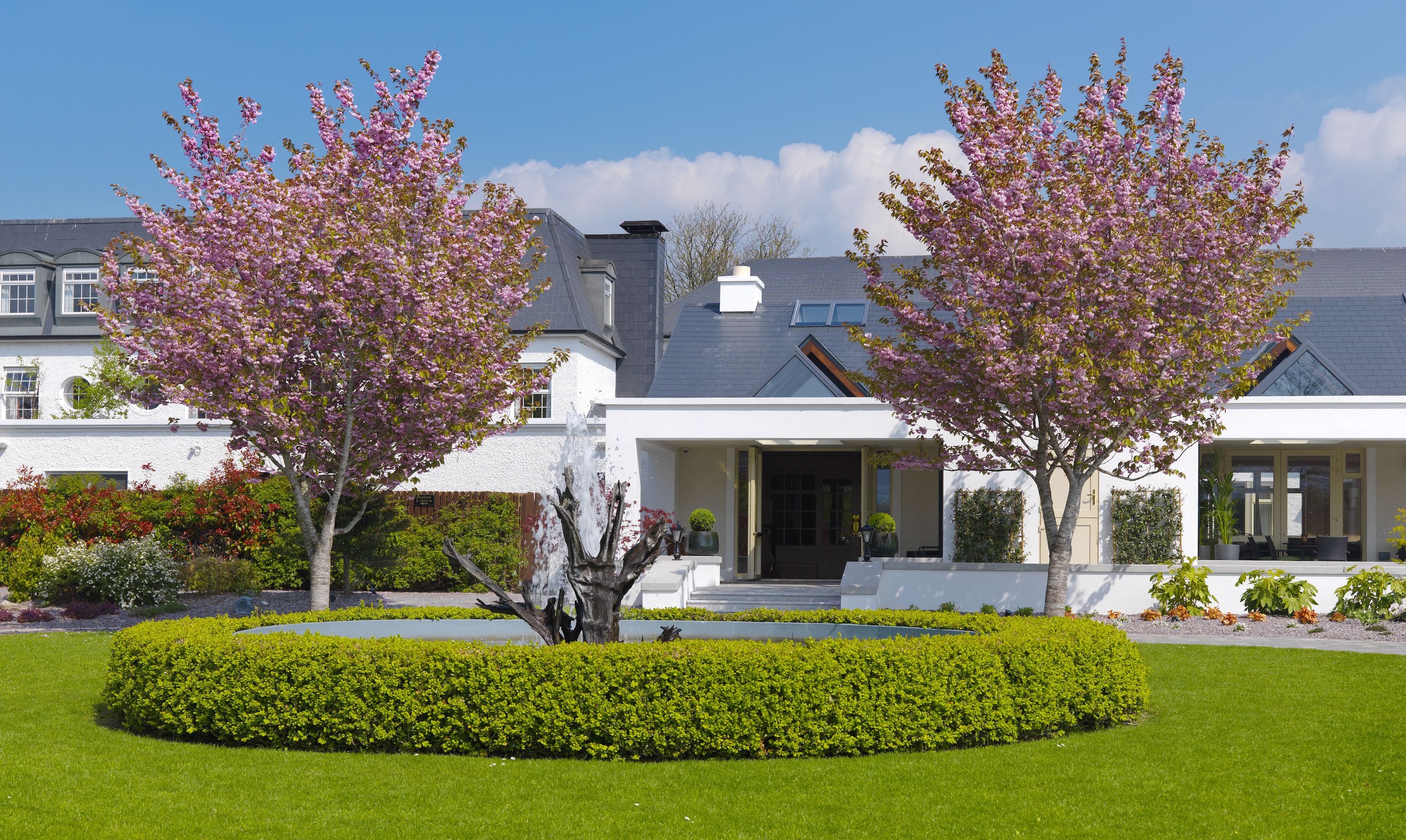 Une maison moderne dotée d’un jardin bien entretenu, comprenant une haie circulaire et des arbres en fleurs.