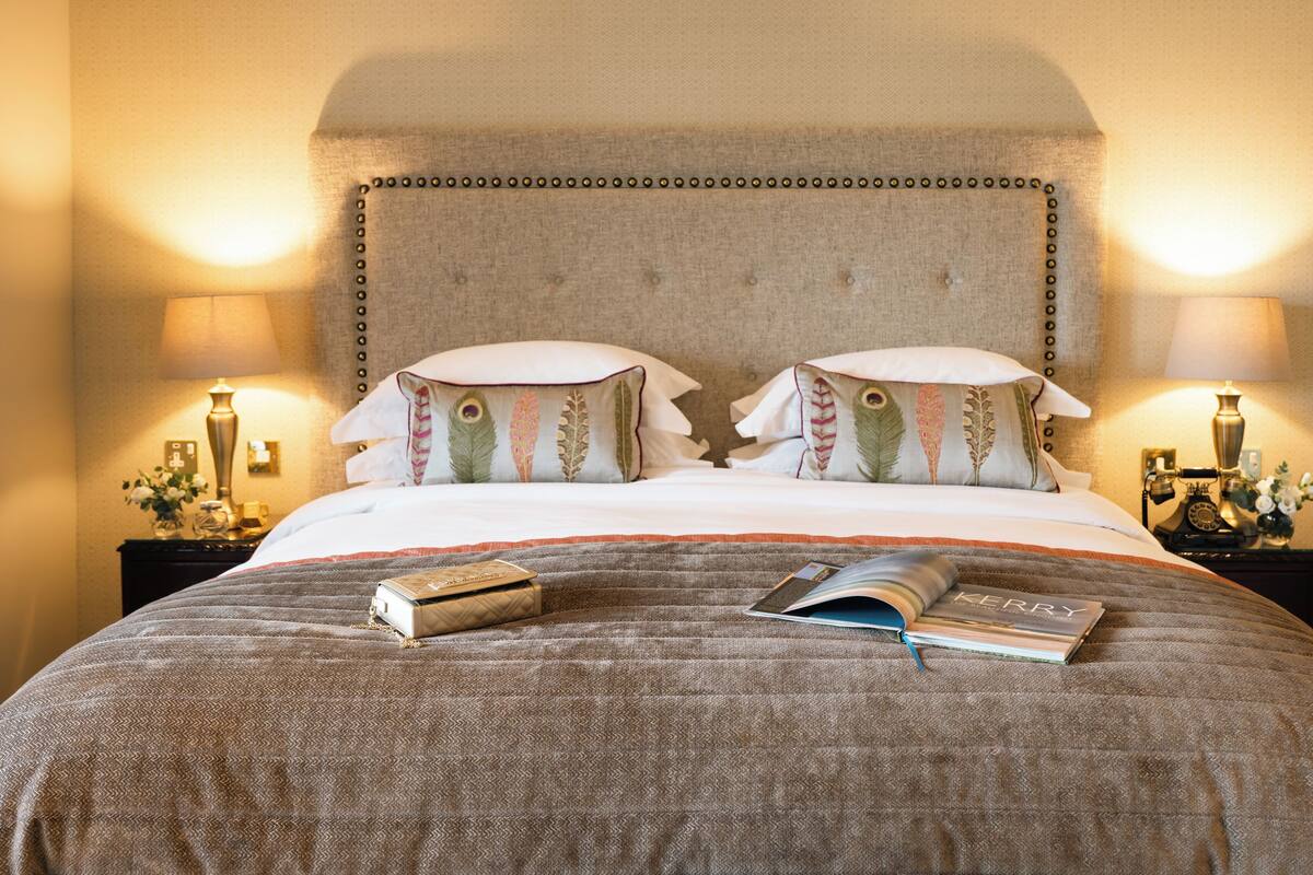 Ein ordentlich gemachtes Bett mit einer gesteppten Decke, zwei Kissen mit Federdesigns, einem Buch und einem Koffer.