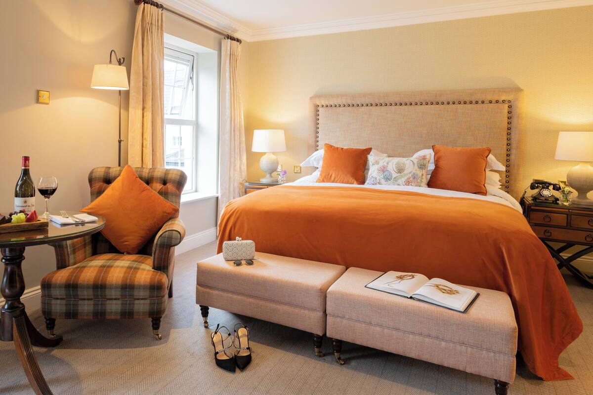 Ein Hotelzimmer mit einem Bett, einem Sessel, einem kleinen Tisch mit Flasche und Glas sowie einer Bank mit Buch.