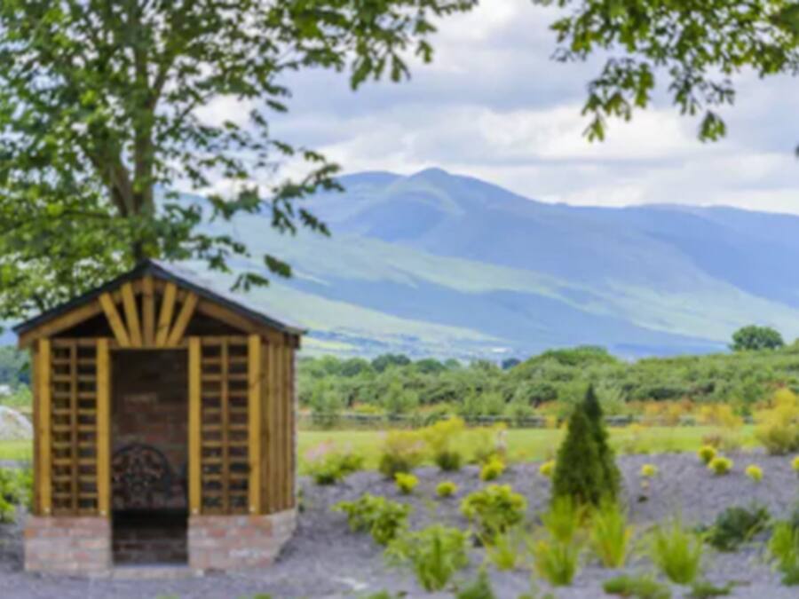 A small wooden structure with a sloped roof, surrounded by greenery and mountains in the background.