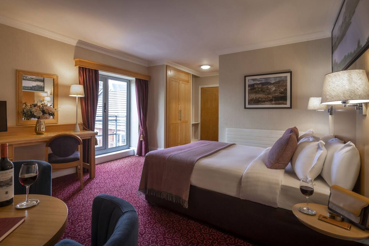 A hotel room with a bed, a desk, a chair, a TV, a window with curtains, and a painting on the wall.