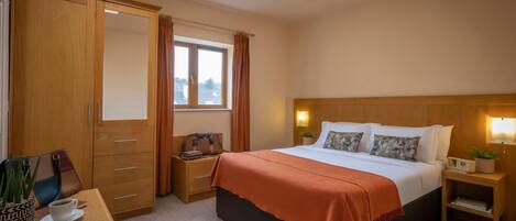 Une chambre d’hôtel comprenant un lit, un bureau avec une chaise, une armoire, une fenêtre avec des rideaux et une table de chevet avec une lampe.