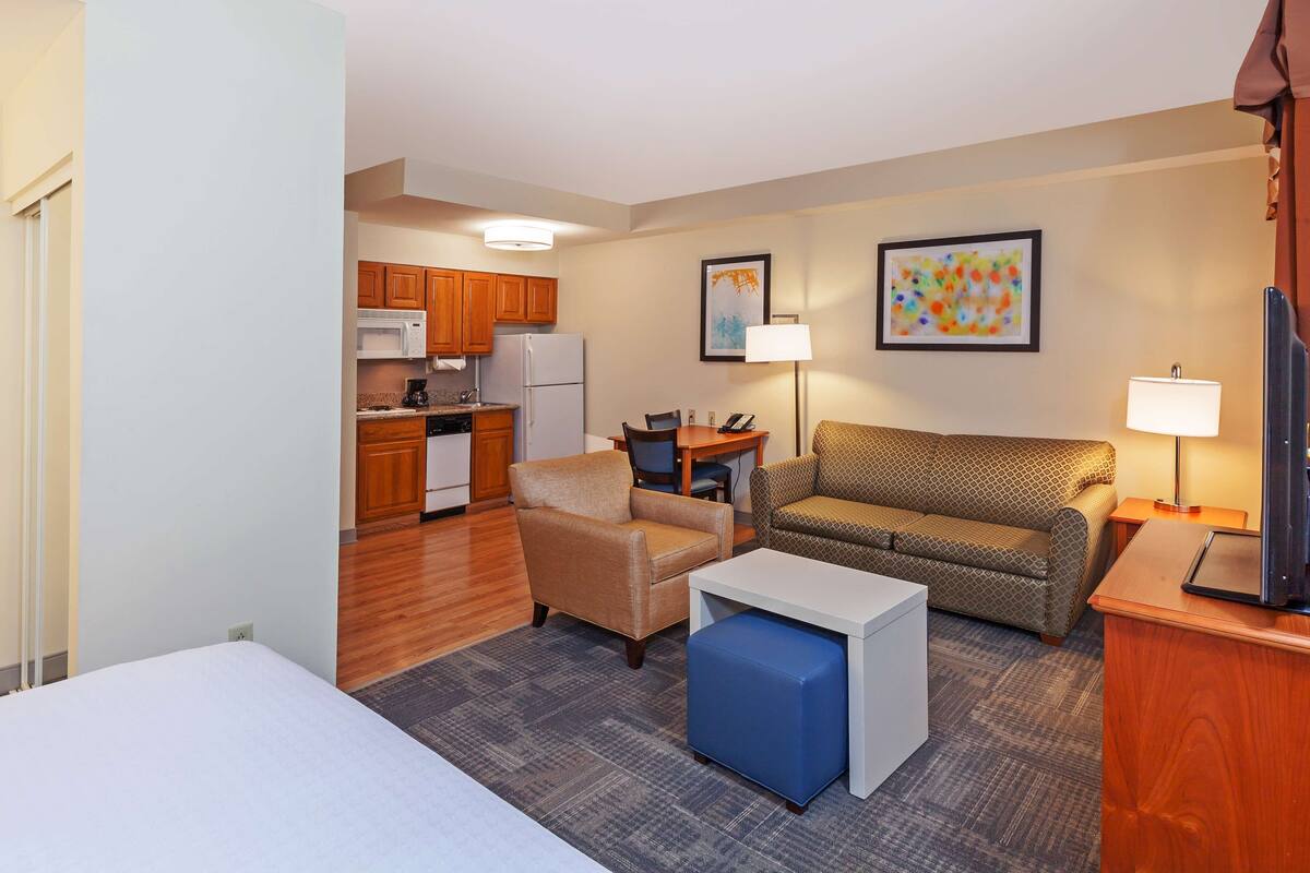 A compact hotel room with a kitchenette, a sofa, a small table, a blue ottoman, and a TV.