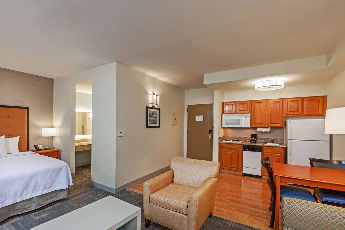 A hotel room with a bed, a chair, a small table, a kitchenette, and a bathroom.