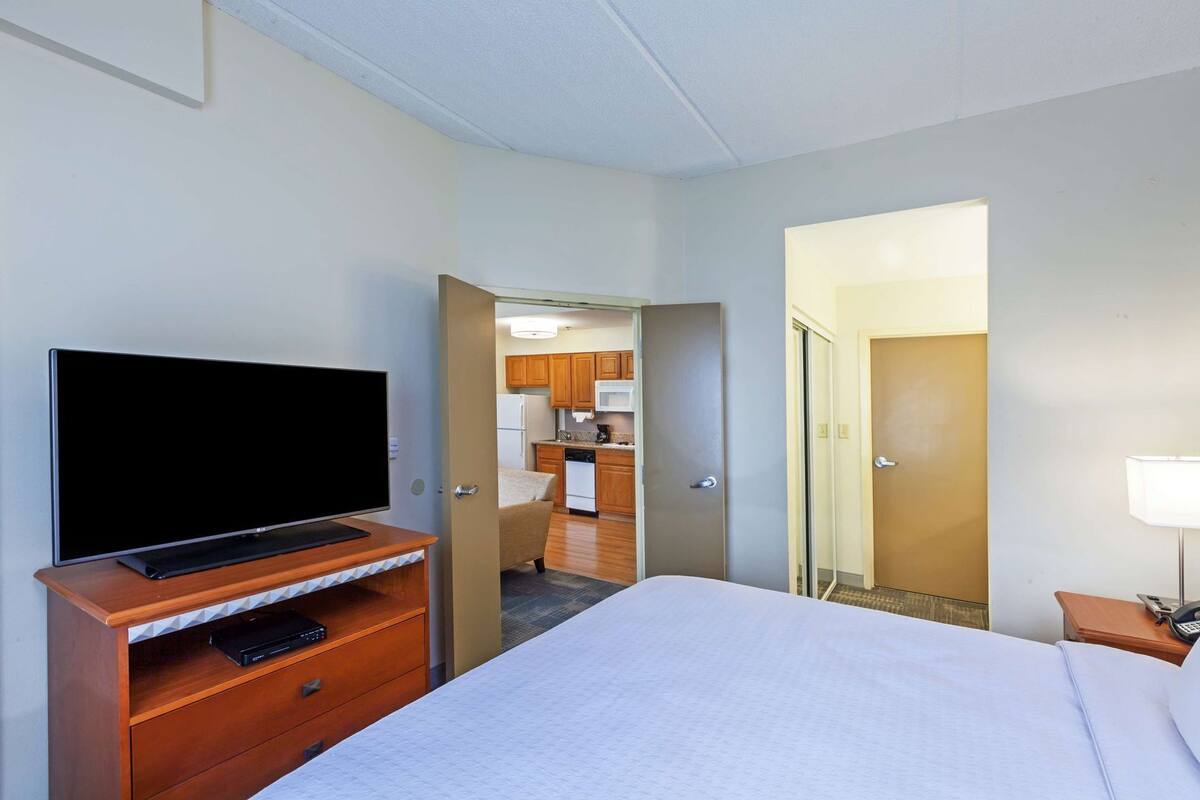 A hotel room with a bed, a TV on a wooden stand, a desk with a lamp, and a view of a kitchen and bathroom through an open door.