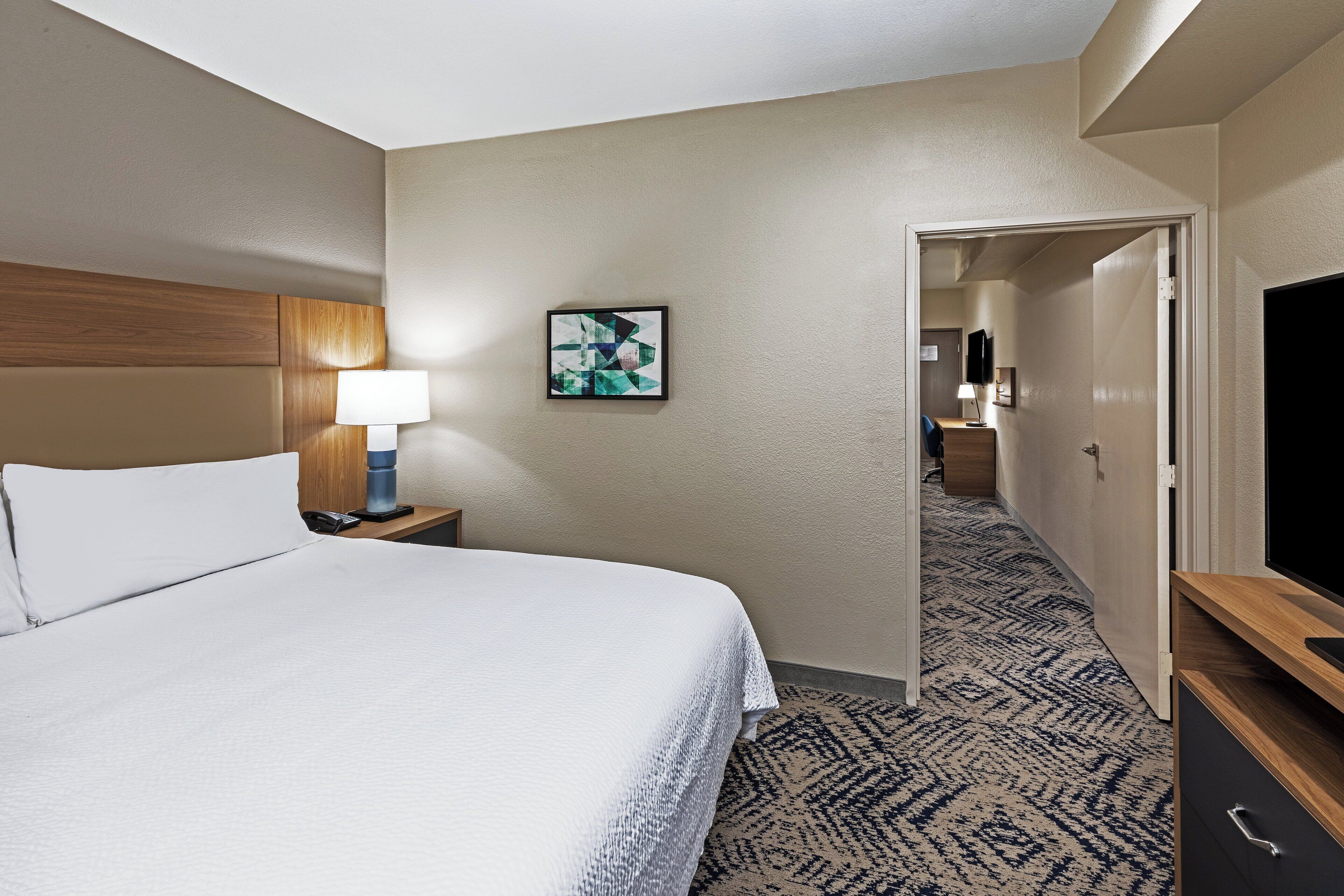 A hotel room with a bed, a nightstand with a lamp, a painting on the wall, and a TV on a wooden stand.